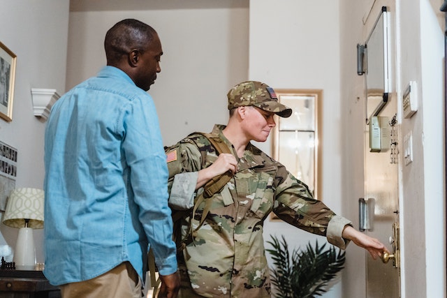 Person in military uniform reaching for their residence's doorknob, their partner standing behind them